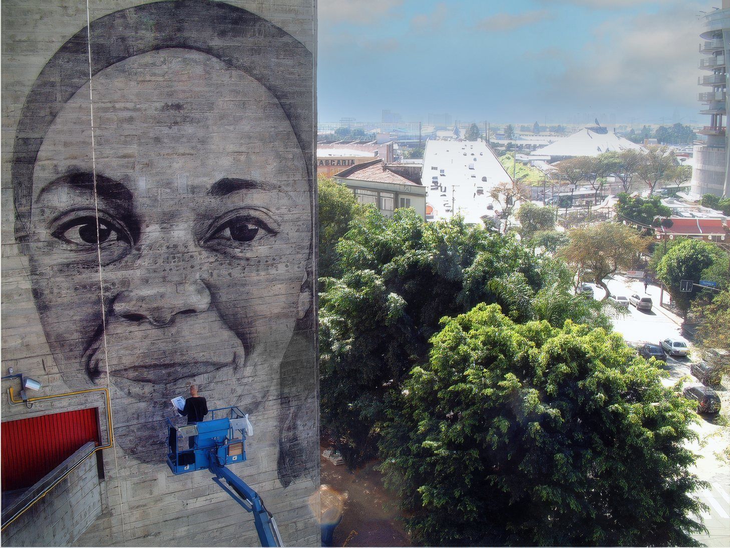 A charcoal portrait drawn across a concrete wall, with the artist working from a lift